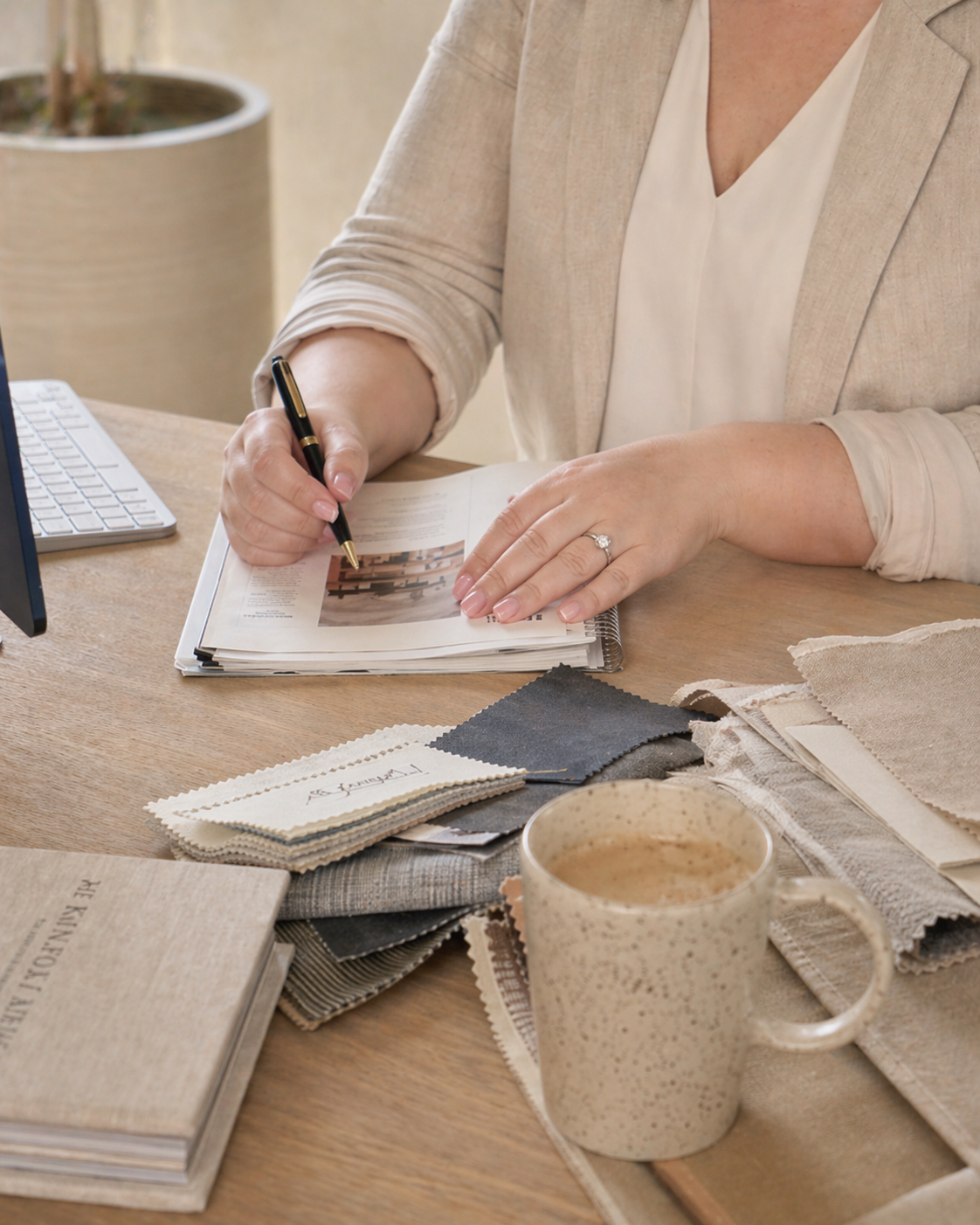Dusty lnen founder in a beige blazer and white top sits at a wooden desk, writing in a sketchbook. The desk is styled with linen fabric swatches, a ceramic mug, and books, suggesting a home decor or interior design context.